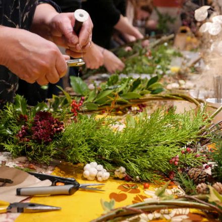 People standing at a table full with green and seasonal foliage being used to decorate rustic Christmas wreaths, at a seasonal workshop. Small craft pliers are being used to cut strands of wire being used to tie leaves, holly, berries, cones and other greenery to the circular base of willow branches. Bowls and vases of berries and dried seed heads are in the middle of the table to choose from.