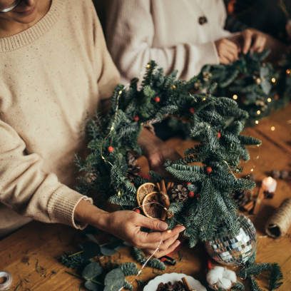 Happy women making Christmas wreath using natural pine branches and festive decorations, standing near wooden table.